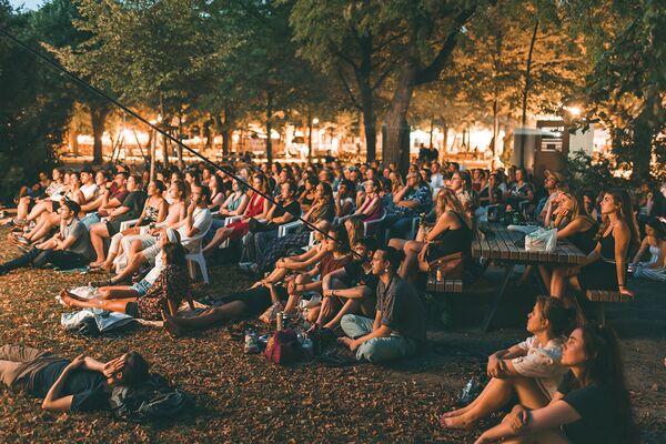 Menschen sitzen am Abend auf dem Boden im Freien und schauen einen Film auf einer großen Leinwand.
