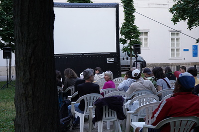 Menschen sitzen am Abend auf dem Boden im Freien und schauen einen Film auf einer großen Leinwand.