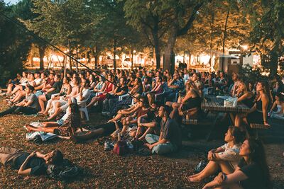 Menschen sitzen am Abend auf dem Boden im Freien und schauen einen Film auf einer großen Leinwand.
